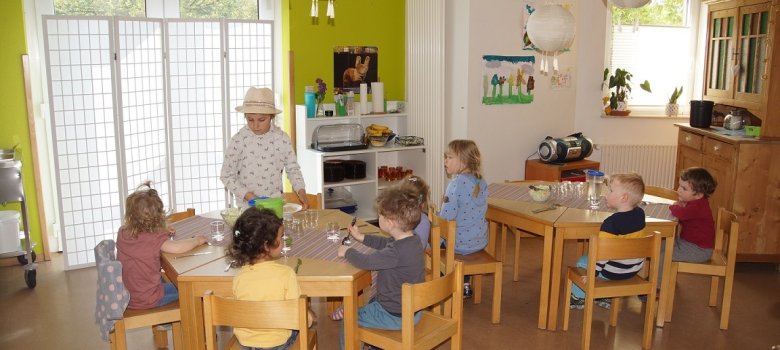 Die Kinder sitzen am Tisch und warten auf das Mittagessen. Ein Mädchen setzt sich mit ihrem Teller hin. 