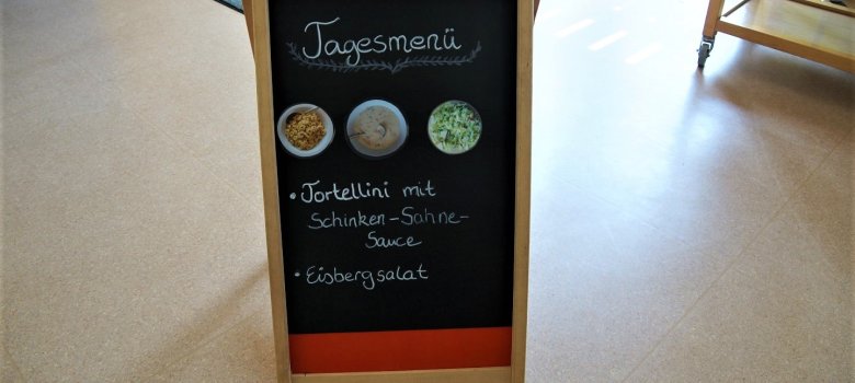 Im Restaurant steht eine Tafel mit dem tagesaktuellen Mittagessen dort steht geschrieben: Tagesmenü Tortellini mit Schinken-Sahne-Sauce Eisbergsalat. In der Mitte der Tafel kleben drei Bilder mit Rortellinis, Salat und der Sauce.