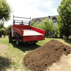 Auf dem Bild sieht man einen roten Kipper auf dem Außengelände der Kita, dessen Ladefläche halb ausgefahren ist. Hinter ihm liegt ein Haufen Boden auf der Wiese den er abgeladen hat. 