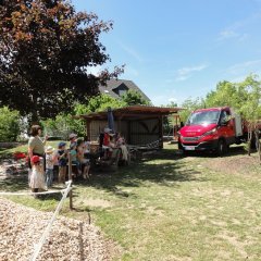 Auf dem Bild sind viele Kinder mit drei Erzieherinnen zu sehen. Sie stehen und sitzen am Tisch und schauen in die selbe Richtung. Dort steht ein rotes Auto mit einem Kipper. 