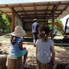 Auf dem Bild sieht man zwei Kinder auf Baumstämmen und ein Kind neben ihnen auf einem Holzbrett. Sie schauen nach vorne auf den Bagger unter der Überdachung der die Betonplatte aufreißt. 