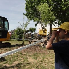 Das Bild zeigt einen Jungen der mit einem roten Fernglas dem Bagger hinter dem Absperrband zuschaut.