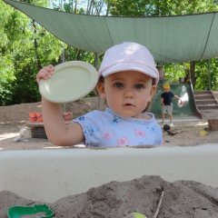 Auf dem Bild sieht man ein kleines Mädchen hinter einer Wanne mit Sand. Sie hält einen Teller nach oben in der Hand und schaut in die Kamera. 