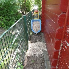 Das Bild zeigt einen Jungen mit einer kleinen Schubkarre gefüllt mit Rindenmulch. Er kippt diesen zwischen dem Zaun und einem Häuschen ab. 