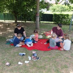 Auf dem Bild sind zwei Erzieher zu sehen, die mit ein paar Kindern auf einer Decke auf der Wiese sitzen. Zwei Kinder liegen mit den Knien und dem Kopf nach unten auf der Decke im Kreis und haben Wäscheklammern an ihrem Shirt befestigt.