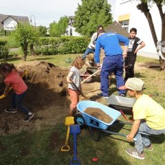 Das Bild zeigt einen großen Erdhügel und viele Kinder und Erwachsene. Alle schaufeln die Erde in Schubkarren. 