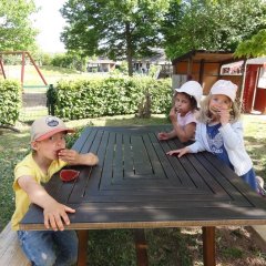 Das Bild zeigt drei Kinder am Tisch die in ein Stück Melone beißen und in die Kamera schauen.