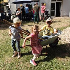 Das Bild zeigt einen Jungen der in einer Schubkarre sitzt und in die Kamera grinst. Neben ihm stehen zwei Mädchen. Im Hintergrund stehen Eltern und Kinder am Essenstand. 