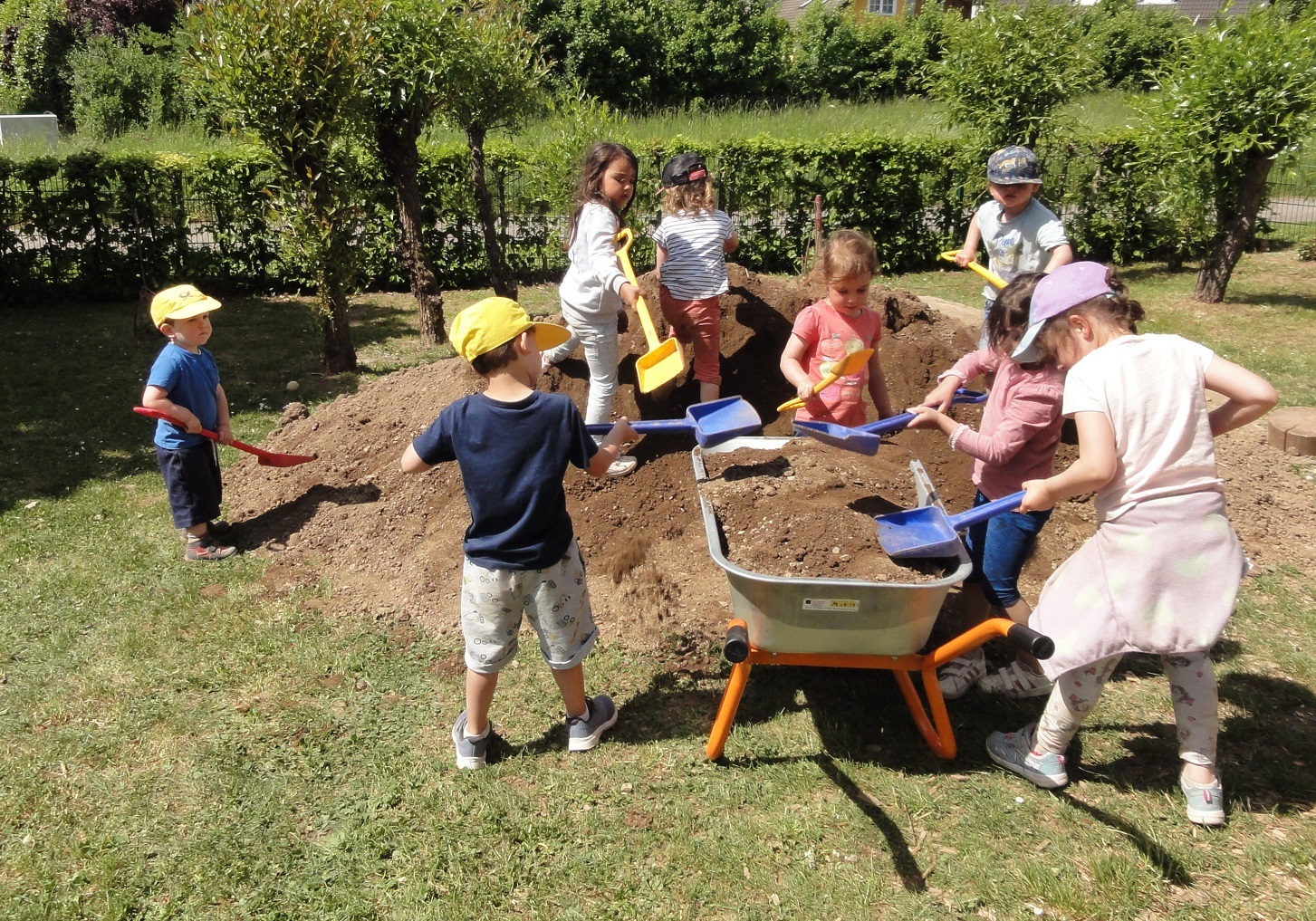 Das Bild zeigt viele Kinder mit Schaufeln die Erde von einem Erdhügel in eine große Schubkarre schaufeln.