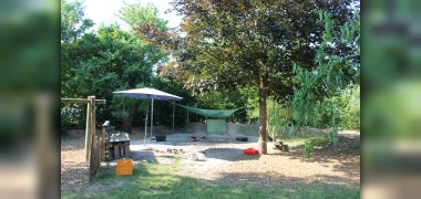 Sandkasten im Garten Auf dem Bild ist ein Sandkasten im Garten der Kita. Daneben steht eine Matschküche und darüber Hängt ein Sonnensegel. In den Sandkasten führt eine Rutsche. Hinter dem Sandkasten ist ein Hügel mit Bäumen.