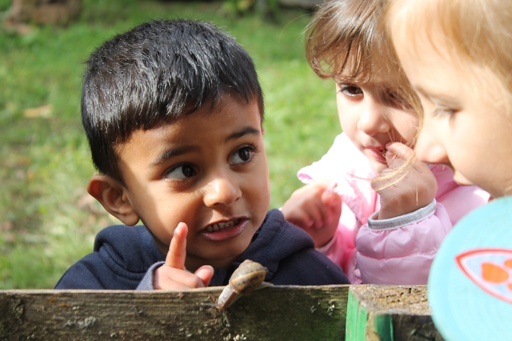 Auf dem Bild sieht man einen kleinen Jungen vor einem Zaun. Neben ihm stehen drei Mädchen und schauen auf eine schwarz gelbe Schnecke auf dem Zaun. Der Junge hat den rechten Zeigefinger erhoben, er schaut und spricht ernst zu den Mädchen.