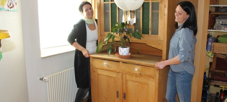 Auf dem Bild sind zwei Erzieherinnen zu sehen, die einen Holzschrank an die Wand schieben. Sie lachen beide. 
