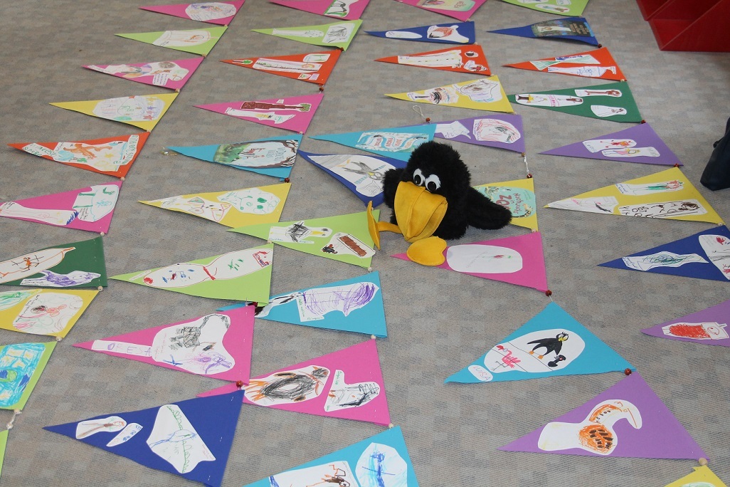 Das Bild zeigt den Raben Rasmus aus der Stadtbücherei in der Mitte von einer langen Girlande mit vielen Wimpeln auf dem Boden. Auf den Wimpeln befinden sich gemalte Bilder der Kinder.