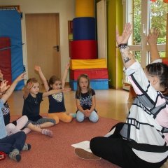 Eine Erzieherin sitzt mit 5 Kindern in einem Kreis und alle haben die Hände in der Luft.