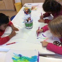2 Kinder sitzen am Tisch und malen die gezeichneten Hände auf weißem Papier bunt aus. 