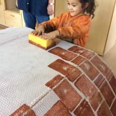 Ein Kind erstellt mit einem Schwamm und brauner Farbe das Muster einer Mauer auf einem großen Pappkarton. 