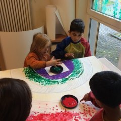 4 Kinder sitzen am Tisch und tupfen mit ihren Fingern kleine Punkte auf einen großen Karton. Auf diesem Karton wurde ein Regenbogen vorgezeichnet. 