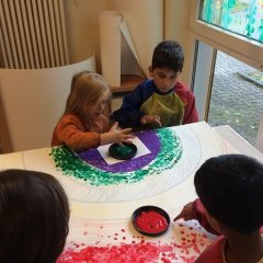 4 Kinder sitzen am Tisch und tupfen mit ihren Fingern kleine Punkte auf einen großen Karton. Auf diesem Karton wurde ein Regenbogen vorgezeichnet. 