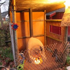 Das Bild zeigt eine kleine Hütte in der ein Lehmbackofen steht mit einem kleinen Zaun davor. Vor der Hütte steht eine Lampe. Dort hängt ein Plakat dran mit den Worten "Wiederverwendung des Lehmbackofens."