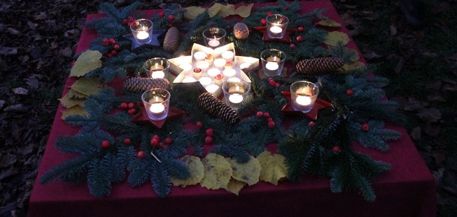 Auf dem Bild ist ein Tisch mit einer roten Tischdecke auf dem Außengelände zu sehen. Auf dem Tisch sind und eine sternförmige Kerze viele Tannenzweige, rote Beeren, Blätter, Gläser mit Teelichtern und Tannenzapfen verteilt. 
