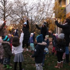 Auf dem Bild sind Kinder und Erwachsene auf dem Außengelände zu sehen. Sie stehen mit Jacken und Mützen in einem Kreis und halten ein LED-Teelicht mit dem Arm nach oben.