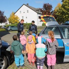 Auf dem Foto sieht man rechts ein Polizeiauto daneben steht ein männlicher Polizist mit Uniform und eine weibliche Polizistin in Uniform. Sie schauen zu den Kindern die vor ihnen stehen und sprechen mit ihnen. Die Kinder schauen ganz gespannt zu den Polizisten. Links neben den Kindern steht die Kita-Leitung und schaut zu den Kindern und Polizisten.