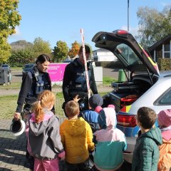 Das Bild zeigt zwei Polizisten in Uniform und vor ihnen stehen viele Kinder und schauen zu ihnen. Neben ihnen ist der Kofferraum des Polizeiauto geöffnet. Der männliche Polizist hält eine hohe Messlatte in rot und weiß in der Hand. Die Polizistin hat eine Polizeimütze in ihrer Hand. 