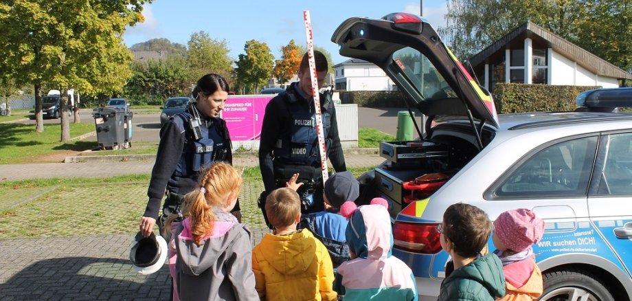 Das Bild zeigt zwei Polizisten in Uniform und vor ihnen stehen viele Kinder und schauen zu ihnen. Neben ihnen ist der Kofferraum des Polizeiauto geöffnet. Der männliche Polizist hält eine hohe Messlatte in rot und weiß in der Hand. Die Polizistin hat eine Polizeimütze in ihrer Hand. 