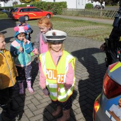 Das Bild zeigt ein Kind das Matschkleidung trägt mit einer Polizei-Warnweste und einer Polizeimütze auf dem Kopf. Das Kind schaut in die Kamera und lächelt.
