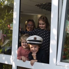 Auf dem Bild sind drei lachende Kinder und eine Erzieherin zu sehen die aus einem geöffneten Fenster schauen. Das vordere Kind hat eine Polizeimütze auf dem Kopf und lacht in die Kamera.