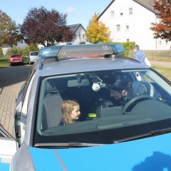 Das Bild zeigt ein Polizeiauto von vorne. Durch die Windschutzscheibe kann man sehen dass auf dem Beifahrersitz ein Kind sitzt neben ihm sitzt eine Polizistin. Sie schaut das Kind an und spricht mit ihm. Beide lachen. 
