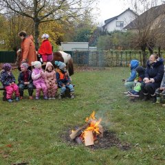 Das Bild zeigt ein paar Kinder die auf dem Außengelände auf Bänken im Kreis sitzen. In der Mitte befindet sich ein Lagerfeuer. Außen um die Kinder geht ein braunes Pony auf dem ein Mädchen sitzt mit einem Reithelm und einem roten Umhang. Das Pferd wird geführt von der Mutter des Kindes.