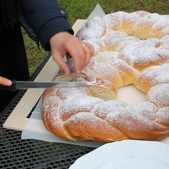 Das Bild zeigt eine riesige geflochtene Martinsbrezel mit Zucker auf einem Tisch. Sie wird von einem langen Messer geschnitten. 