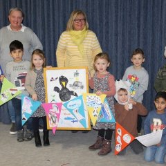 Das Bild zeigt 9 Kinder und zwei Erzieherinnen die in zwei Reihen vor einem blauen Vorhang stehen. Sie halten gemeinsam eine Girlande mit Wimpeln und einem Bilderrahmen mit einem selbst gemalten Bild des Raben Rasmus in der Hand. 