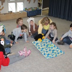 Das Bild zeigt neun Kinder und zwei Erzieherinnen die in einem Kreis auf dem Teppich sitzen. Eine Erzieherin hat den Raben als Handpuppe auf der Hand und im Schnabel des Raben steckt ein langes dünnes Geschenk in blauem Geschenkpapier. Die Kinder schauen zu dem Geschenk.