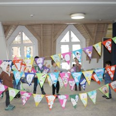 Das Bild zeigt eine lange Girlande mit vielen Wimpel die von den Erziehern und Kindern festgehalten wird. Auf den Wimpeln befinden sich Bilder die die Kinder zu den Bilderbüchern gemalt haben, die der Rabe Rasmus mit in die Kita gebracht hat.