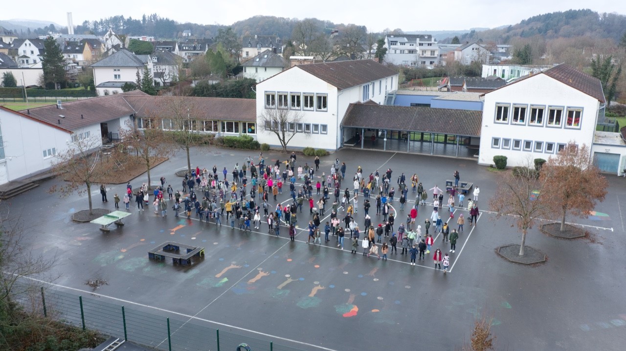 Das Bild zeigt die GMGS aus der Vogelperspektive. Auf dem Schulhof stehen alle Kinder.
